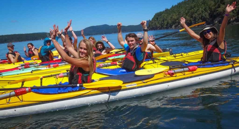 a group of students in kayaks raise their hands in celebration on an outward bound course